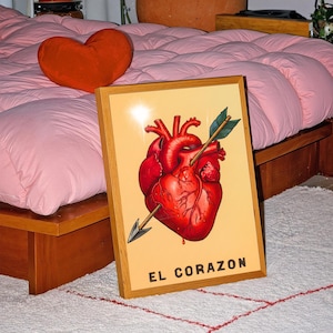 May include: A rectangular framed print featuring a detailed illustration of a red anatomical heart pierced by an arrow, set against a light beige background. The text EL CORAZON is printed in black capital letters at the bottom of the artwork.