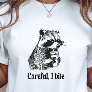 May include: White t-shirt featuring a black and white illustration of a raccoon holding a beverage. The text "Careful, I bite" is printed below the raccoon. The shirt is a casual style.