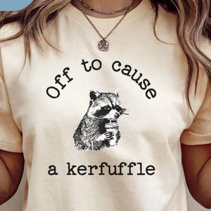May include: Cream-colored t-shirt with the text "Off to cause a kerfuffle" and a black and white illustration of a raccoon holding a drink. The word "Natural" is in a blue box in the upper left corner.