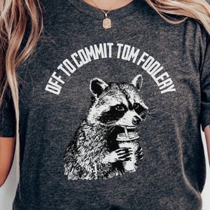 May include: Dark grey heather t-shirt with a raccoon illustration holding a coffee cup. The text "OFF TO COMMIT TOM FOOLERY" is arched above the raccoon. The shirt is a dark grey colour.