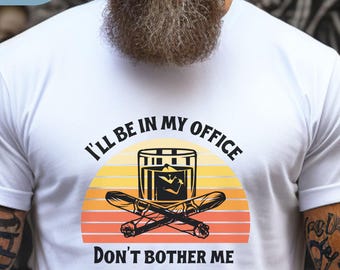 May include: White t-shirt with a graphic design featuring a whiskey glass, crossed cigars, and the text "I'll be in my office, Don't bother me." The design includes a sunset-style background in orange and yellow.