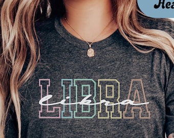 May include: Dark grey heather t-shirt with the word "LIBRA" in a colorful, outlined font. The word "libra" is written in a cursive font over the outlined word. The shirt is a crew neck style.