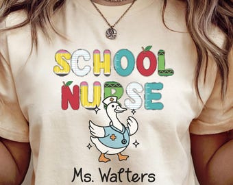 May include: A light beige t-shirt with the words "SCHOOL NURSE" in colorful letters and a cartoon duck wearing a nurse's uniform. The name "Ms. Walters" is printed below the duck.