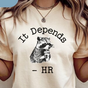 May include: Cream-colored t-shirt with the text "It Depends - HR" and a black and white illustration of a raccoon holding a cup. The word "Natural" is in a blue box in the upper left corner.