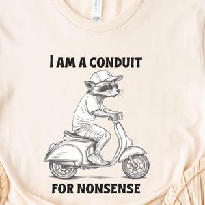 May include: A beige t-shirt with a black and white illustration of a raccoon wearing sunglasses and riding a scooter. The text "I am a conduit for nonsense" is printed on the shirt.