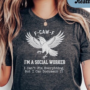 May include: Dark grey heather t-shirt with a white graphic of a raven and the text "F-CAW-F" above it. Below the raven, the text reads "I'M A SOCIAL WORKER" and "I Can't Fix Everything, But I Can Document It."