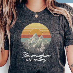 May include: Dark grey heather t-shirt featuring a circular graphic of mountains, trees, and a sunset. The text "The mountains are calling" is below the graphic. The shirt is a short-sleeved crew neck.