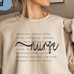 May include: Sand-colored sweatshirt with the word "nurse" in a cursive font. The sweatshirt also features a list of nursing-related terms such as "scrubs," "vital signs," and "teamwork."