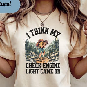 May include: Cream-colored t-shirt with the text "I THINK MY CHECK ENGINE LIGHT CAME ON" and a graphic of a hiker in a mountain scene. The shirt has short sleeves and a crew neck.