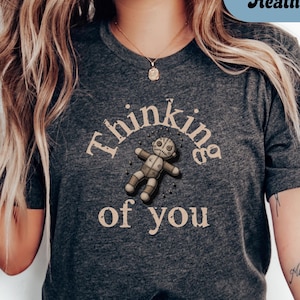 May include: Dark grey heather t-shirt with the text "Thinking of you" and a voodoo doll graphic. The shirt is a dark grey color and has a crew neck. The graphic is in a vintage style.