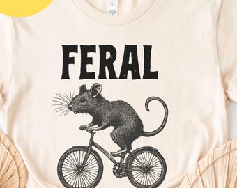May include: A beige t-shirt with the word "FERAL" printed in black. A black and white illustration of a rat riding a bicycle is below the word.