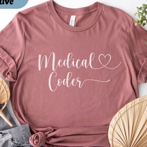 May include: Heather mauve t-shirt with the words "Medical Coder" in white script, featuring a heart symbol. The shirt is styled with rolled-up sleeves and a knot at the waist. Decorative fans and greenery are in the background.