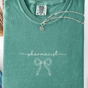 May include: A teal Comfort Colors t-shirt with the word "pharmacist" in cursive and a bow graphic. A silver necklace with a circular pendant is draped across the shirt. The shirt tag reads M-M-M.