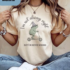 May include: Cream-colored t-shirt with the text "I'm not always right, but I'm never wrong." A cartoon frog wearing a wizard hat rides a scooter. The word "Natural" is in a blue box in the upper left corner.