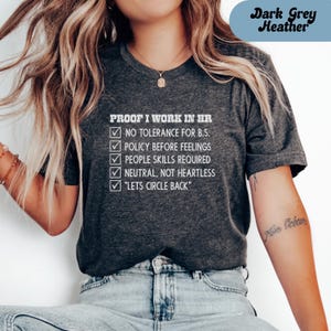 May include: Dark grey heather t-shirt with white text that reads "PROOF I WORK IN HR" followed by a list of HR-related statements. The shirt also has the text "Dark Grey Heather" in a blue oval.