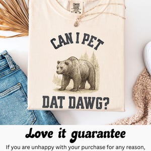 May include: Ivory-colored t-shirt with the text "CAN I PET DAT DAWG?" and a graphic of a bear. The shirt is made of a soft material and has a crew neck. A gold necklace with a pendant is draped on the shirt.