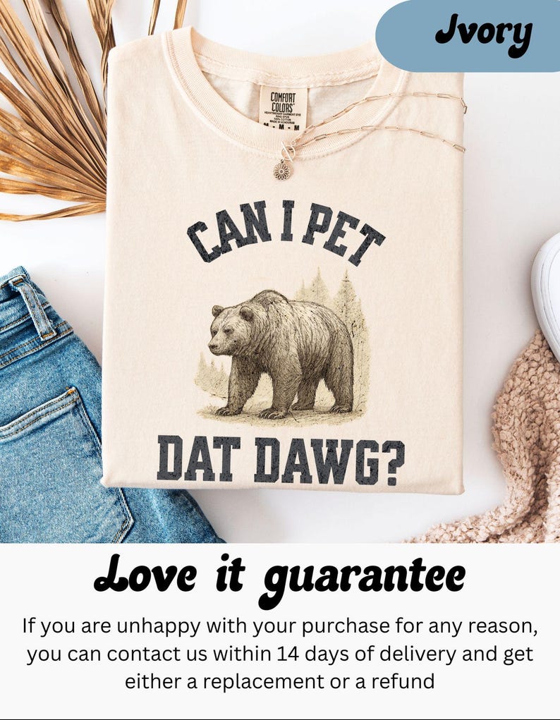 May include: Ivory-colored t-shirt with the text "CAN I PET DAT DAWG?" and a graphic of a bear. The shirt is made of a soft material and has a crew neck. A gold necklace with a pendant is draped on the shirt.