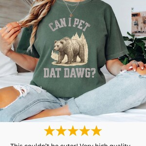May include: Moss green t-shirt with the phrase "CAN I PET DAT DAWG?" and a graphic of a bear. The shirt is a relaxed fit and has a vintage style. The text and bear graphic are in a distressed style.
