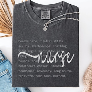May include: A dark gray t-shirt with the word "nurse" in a cursive font. The shirt features a list of nursing-related terms in a light gray font. A silver necklace with a pendant is on the shirt.
