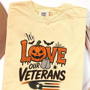 May include: A butter yellow t-shirt featuring a graphic with the text We LOVE OUR VETERANS. The design includes a jack-o'-lantern, two silver dog tags, bats, a spider web, a spider, and a stylised American flag with orange and black stripes.