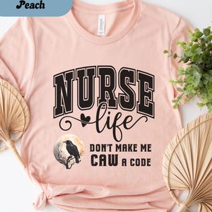 May include: Heather peach t-shirt with the words "NURSE life" in black block letters. Below the words is a graphic of a raven on a branch in front of a full moon. The text "DON'T MAKE ME CAW A CODE" is below the graphic.
