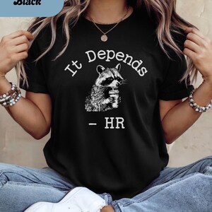 May include: Black t-shirt with the text "It Depends" above a raccoon illustration holding a cup. Below the raccoon is the text "- HR". The shirt has a crew neck.