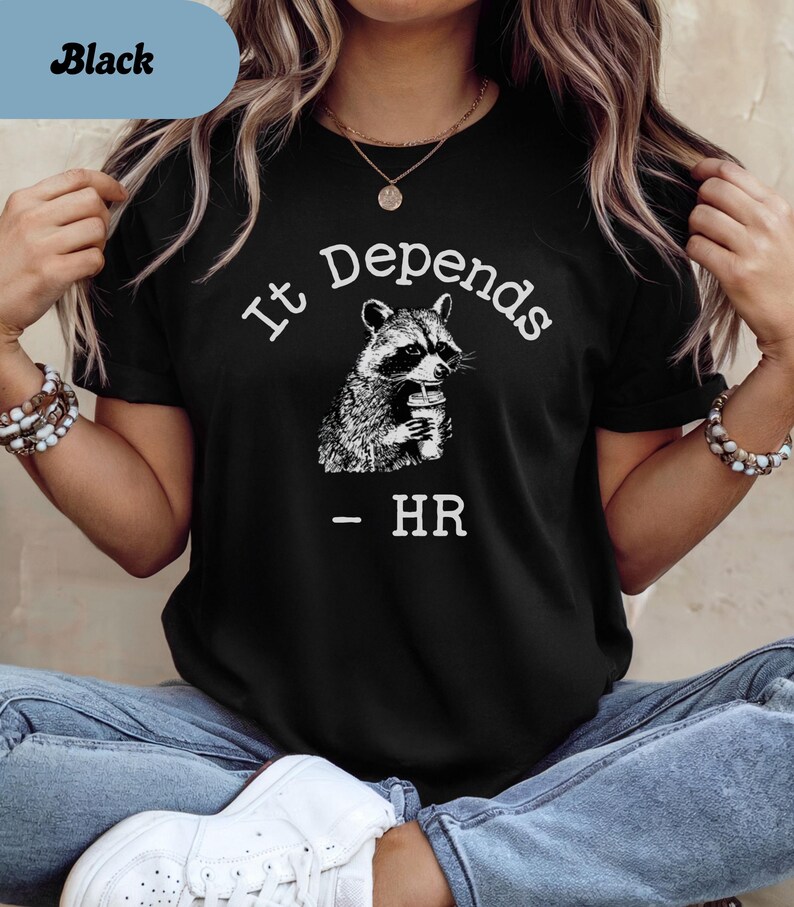 May include: Black t-shirt with the text "It Depends" above a raccoon illustration holding a cup. Below the raccoon is the text "- HR". The shirt has a crew neck.