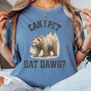 May include: Blue jean colored t-shirt with the text "CAN I PET DAT DAWG?" above a graphic of a bear. The bear is brown and tan, and the text is in a dark gray color. The shirt is a solid color.
