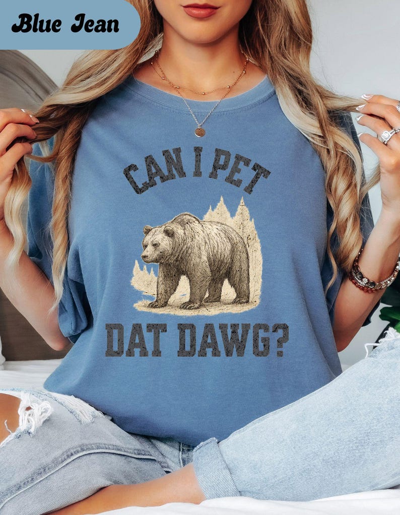 May include: Blue jean colored t-shirt with the text "CAN I PET DAT DAWG?" above a graphic of a bear. The bear is brown and tan, and the text is in a dark gray color. The shirt is a solid color.
