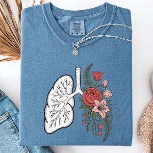 Peut inclure: T-shirt bleu jean avec un motif de poumons et de fleurs. Les poumons sont blancs avec des contours noirs, et les fleurs sont rouges et roses. Un collier argenté avec un pendentif soleil est sur le t-shirt.