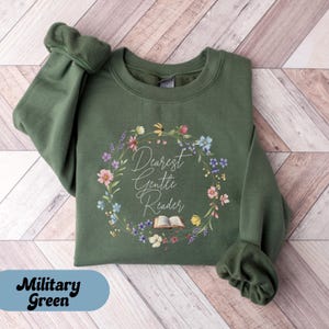 May include: Military green sweatshirt with the text "Dearest Gentle Reader" inside a floral wreath. The design includes an open book. The sweatshirt is folded on a light wood-grain surface.
