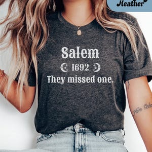 May include: Dark grey heather t-shirt with the text "Salem 1692 They missed one." The text is in white, with crescent moon graphics. The shirt is a dark grey color.