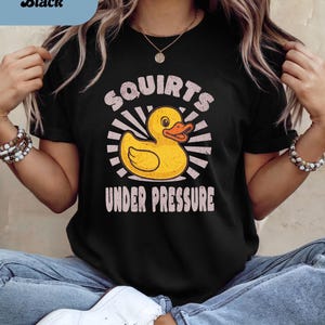 以下が含まれることがあります： 黄色いアヒルのグラフィックと「Squirts Under Pressure」の文字が入った黒いTシャツ。アヒルはオレンジ色のくちばしを持ち、白い光線に囲まれています。カジュアルな普段着です。