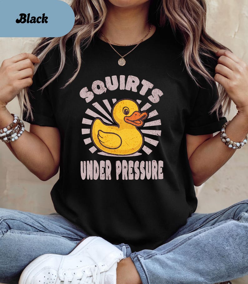 以下が含まれることがあります： 黄色いアヒルのグラフィックと「Squirts Under Pressure」の文字が入った黒いTシャツ。アヒルはオレンジ色のくちばしを持ち、白い光線に囲まれています。カジュアルな普段着です。