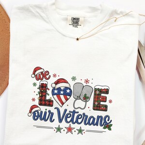 May include: A white t-shirt featuring a graphic that reads "we LOVE our Veterans" in red, blue, and plaid lettering. The design includes a heart with an American flag pattern, two silver dog tags, Santa hats, snowflakes, and holly accents.
