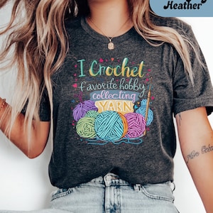 May include: Dark grey heather t-shirt with the text "I Crochet favorite hobby collecting YARN" in colorful lettering. The design includes illustrations of yarn balls and a crochet hook, perfect for crafters and yarn enthusiasts.