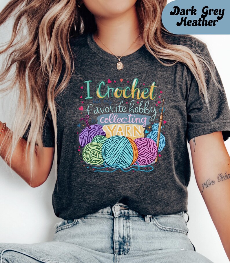 May include: Dark grey heather t-shirt with the text "I Crochet favorite hobby collecting YARN" in colorful lettering. The design includes illustrations of yarn balls and a crochet hook, perfect for crafters and yarn enthusiasts.
