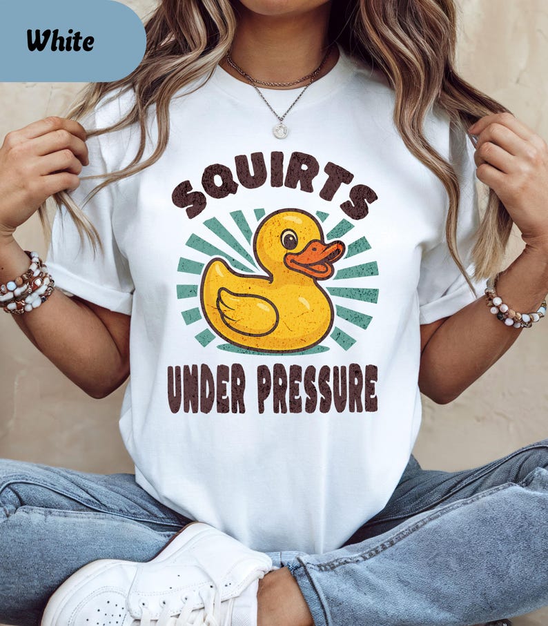 以下が含まれることがあります： 黄色いアヒルのイラストがプリントされた白いTシャツ。オレンジ色のくちばしと「Squirts Under Pressure」という茶色の文字が特徴です。アヒルは、ターコイズと白のバーストデザインを背景にしています。