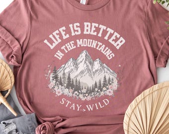 May include: Mauve t-shirt with the text "Life is Better in the Mountains, Stay Wild." The graphic features a mountain range, trees, and floral accents. The shirt is made of a soft, comfortable material.