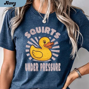 以下が含まれることがあります： オレンジ色のくちばしを持つ黄色のラバーダックのグラフィックと「Squirts Under Pressure」のテキストが特徴のヘザーネイビーTシャツ。シャツの上部には「Heather Navy」の文字も入っています。