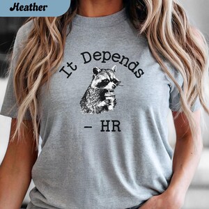 May include: Athletic heather gray t-shirt with the text "It Depends - HR" and a black and white illustration of a raccoon holding a beverage. The shirt is a short-sleeved crew neck.