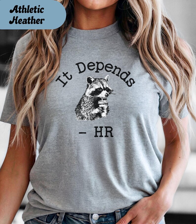 May include: Athletic heather gray t-shirt with the text "It Depends - HR" and a black and white illustration of a raccoon holding a beverage. The shirt is a short-sleeved crew neck.