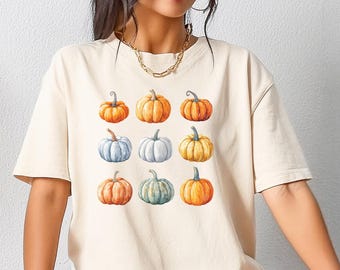 May include: A cream colored t-shirt with a pattern of nine watercolor pumpkins in various shades of orange, yellow, white, and blue.