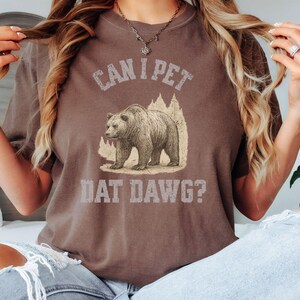 May include: Espresso-colored t-shirt with the text "CAN I PET DAT DAWG?" above a bear graphic. The bear is standing in front of a mountain and tree design. The shirt is a crew neck and has short sleeves.