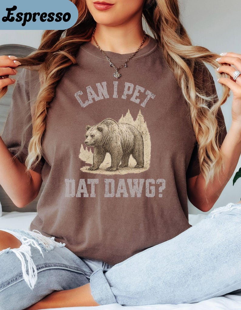 May include: Espresso-colored t-shirt with the text "CAN I PET DAT DAWG?" above a bear graphic. The bear is standing in front of a mountain and tree design. The shirt is a crew neck and has short sleeves.