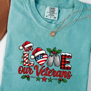 May include: A sea foam green t-shirt featuring a graphic that reads "we LOVE our Veterans". The design includes a candy cane letter L, a heart with an American flag pattern, silver dog tags, and a plaid letter E, all topped with Santa hats.