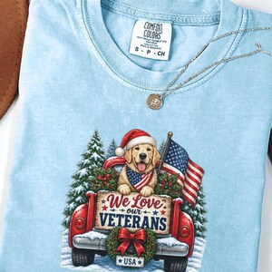 May include: A light blue t-shirt featuring a graphic of a golden retriever wearing a Santa hat and a flag bandana in the back of a red truck. The truck bed displays a sign that reads We Love our VETERANS with a wreath and a USA license plate.