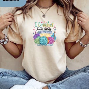 May include: A light beige t-shirt with the text "I Crochet but my favorite hobby is collecting YARN" and colorful yarn balls. The word "Natural" is in a blue rectangle in the upper left corner.