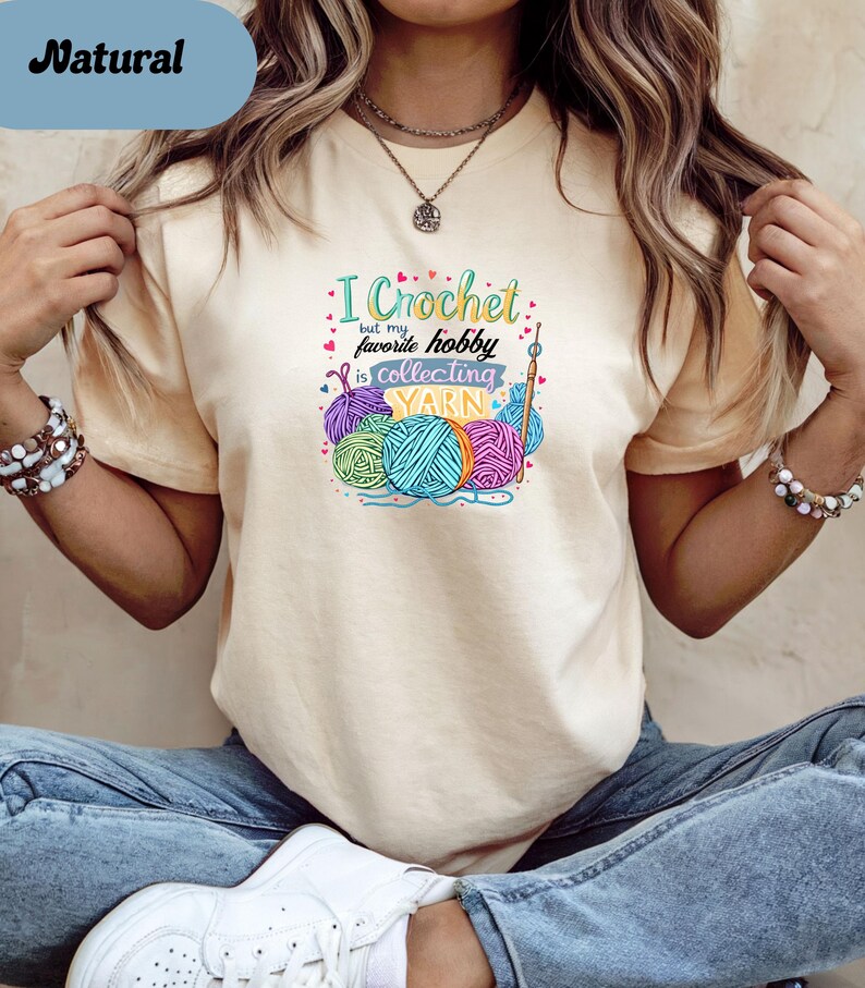 May include: A light beige t-shirt with the text "I Crochet but my favorite hobby is collecting YARN" and colorful yarn balls. The word "Natural" is in a blue rectangle in the upper left corner.