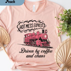 May include: Heather peach t-shirt with a pink train graphic and the text "HOT MESS EXPRESS" above the train. Below the train, the text reads "Driven by coffee and chaos".